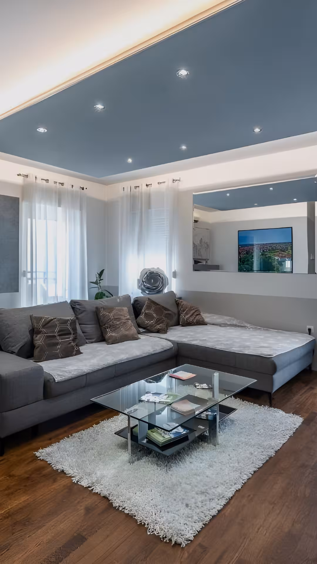 Modern living room with gray sectional sofa, glass coffee table on white rug, wooden floor, and blue accent ceiling with recessed lights.